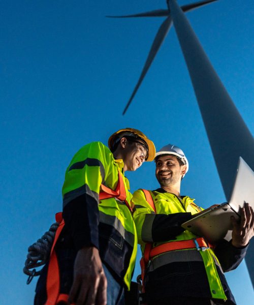 Diverse ethnicity male technicians working in the wind turbines field. Innovation engineer men use laptop computer work to develop environmental renewable clean energy outdoors at windmill power farm.